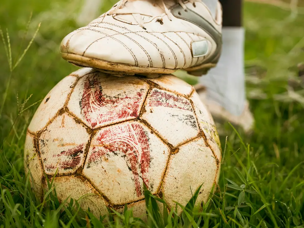 A worn football boot resting on top of a scuffed football on grass